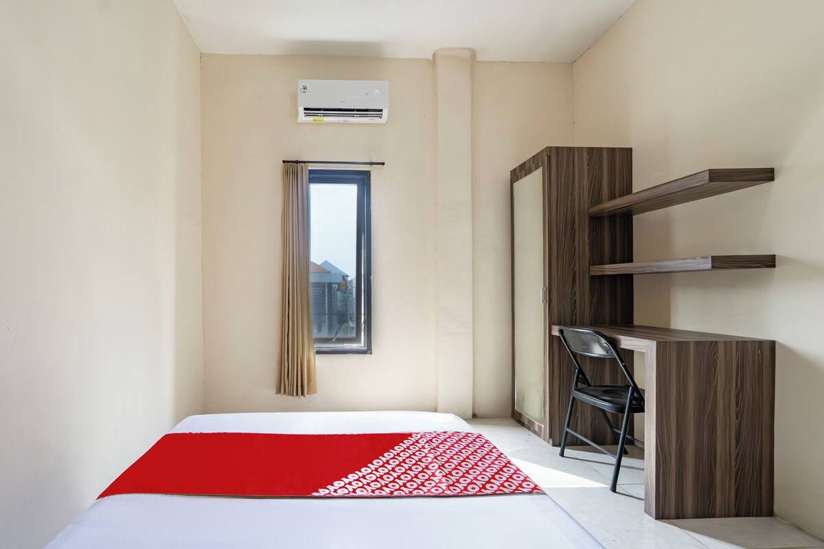 A hotel room with a bed, a desk with a chair, an air conditioner, a window with curtains, and a view of buildings outside.