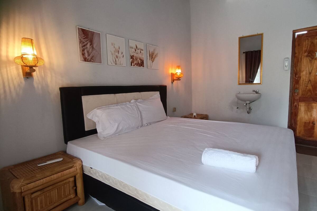 A neatly made bed with white linens, a wooden nightstand, a wall-mounted sink, and framed botanical prints on the wall.