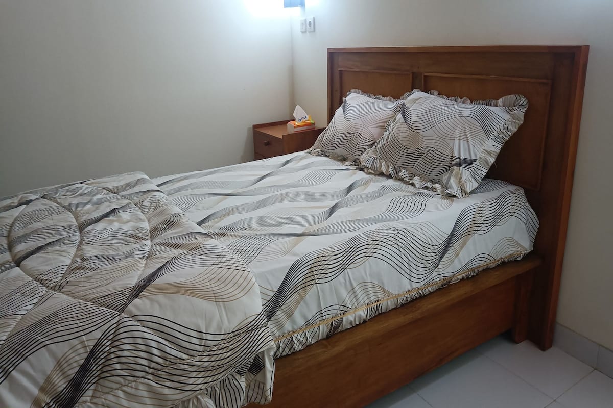 Una cama bien hecha con una colcha estampada y almohadas, un cabecero de madera y una mesita de noche con una caja de pañuelos.