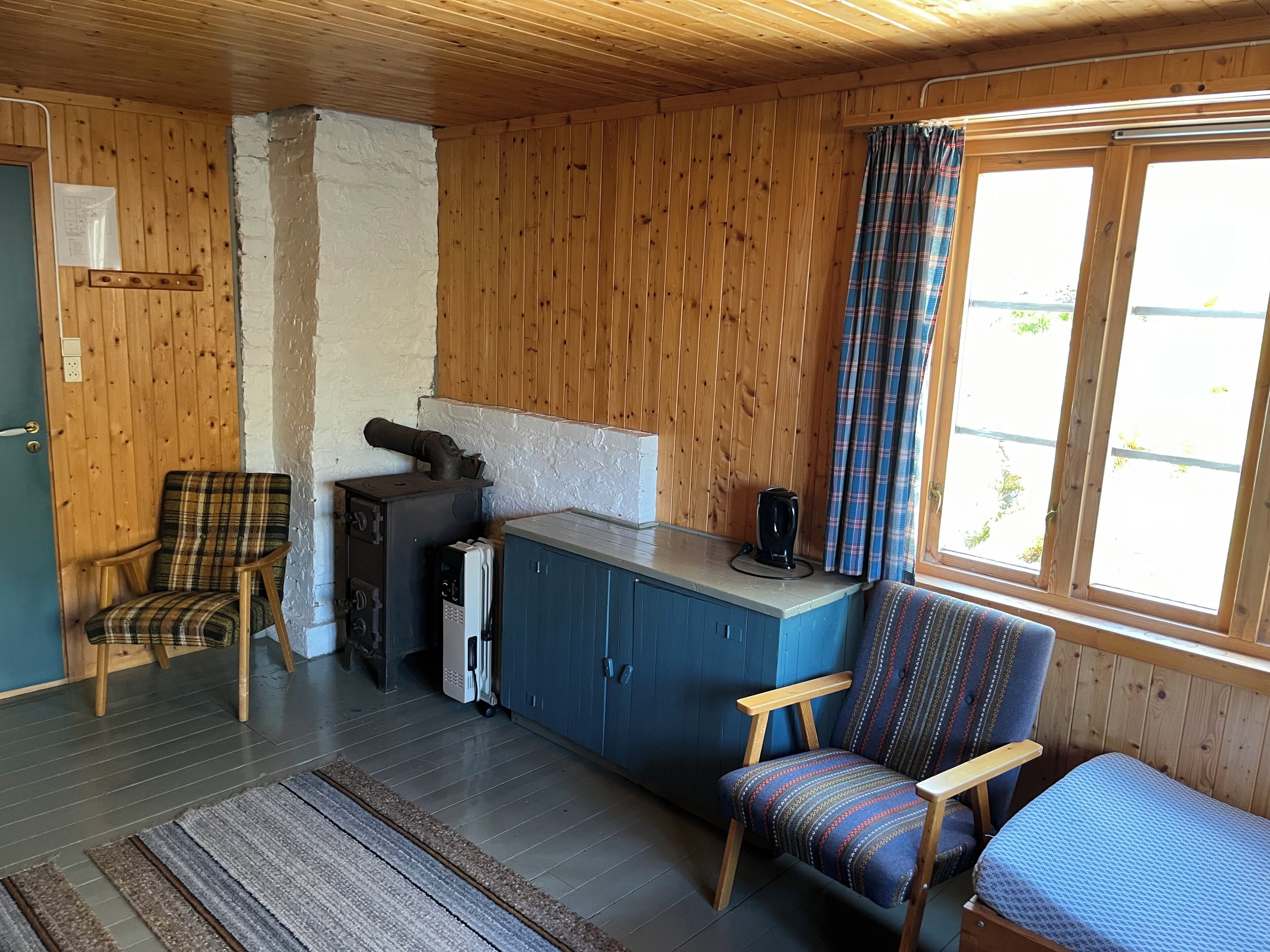 A cozy cabin interior with wooden walls, a kitchenette, a plaid-patterned chair, a blue cabinet, and a window with plaid curtains.