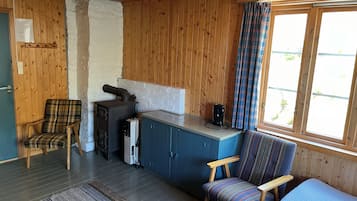A cozy cabin interior with wooden walls, a kitchenette, a plaid-patterned chair, a blue cabinet, and a window with plaid curtains.