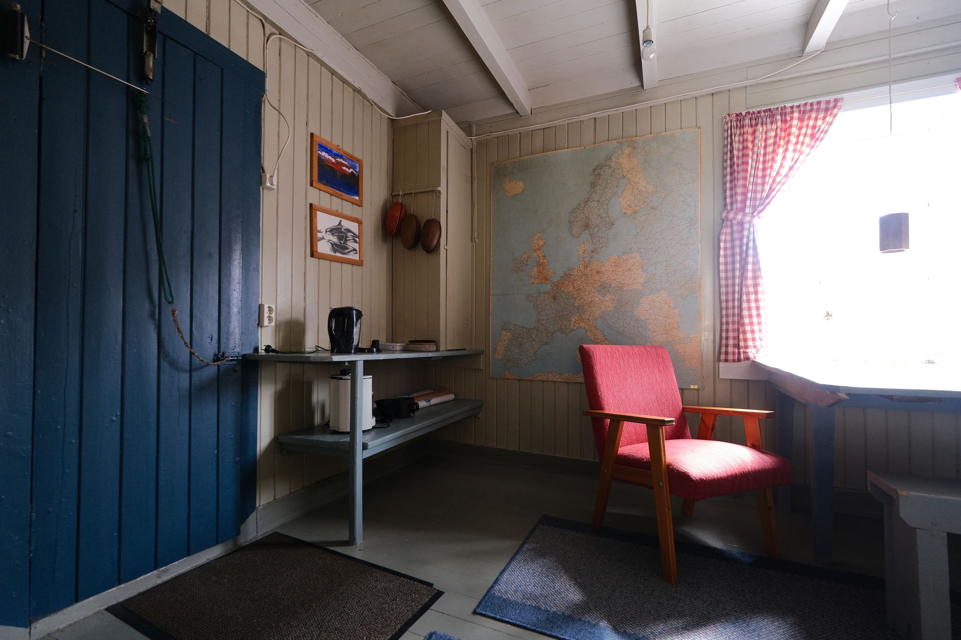 Una habitación con puerta azul, un mapa en la pared, una silla roja y una ventana con cortina a cuadros.