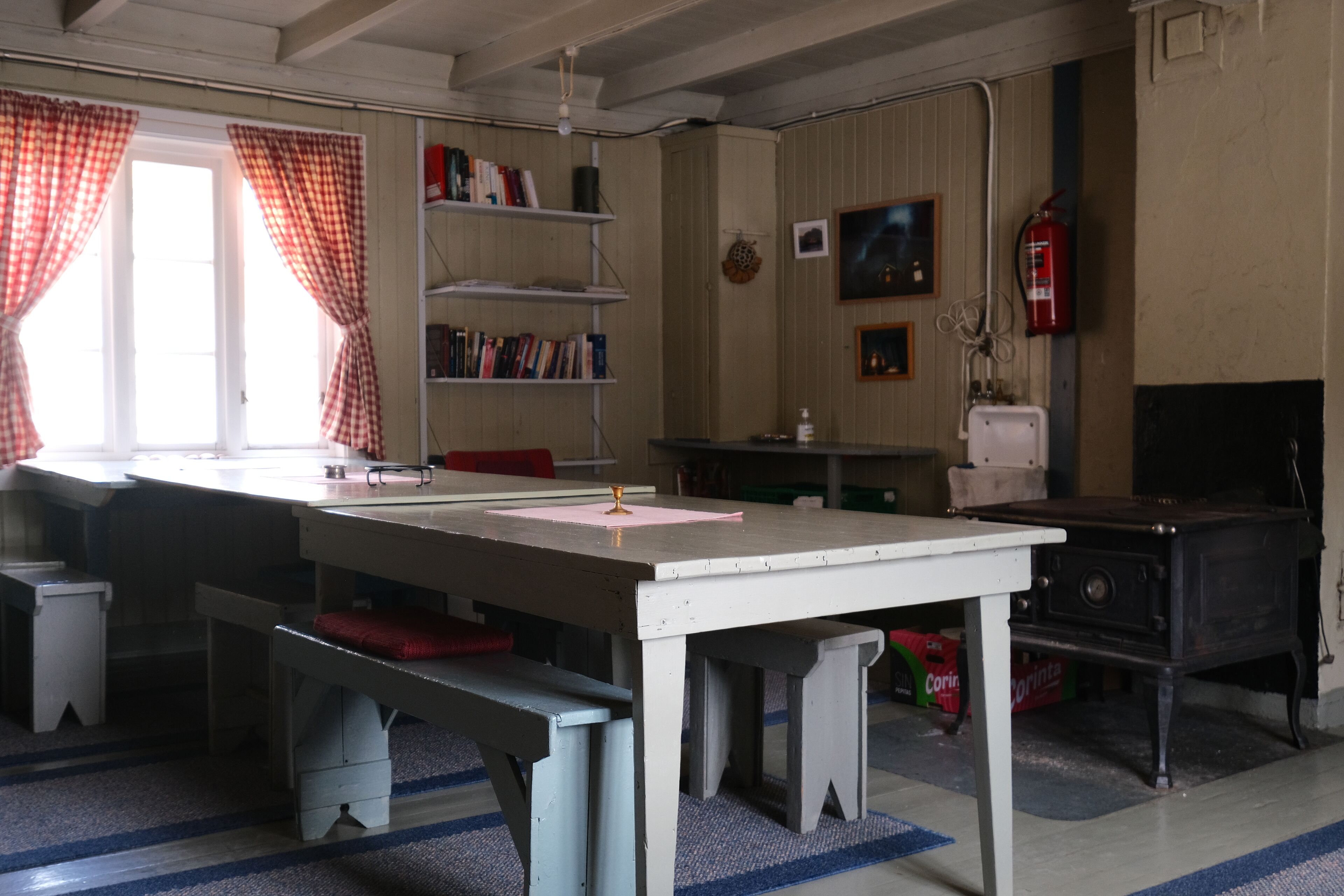 A room with a long table, benches, a bookshelf, a fire extinguisher, and a stove.