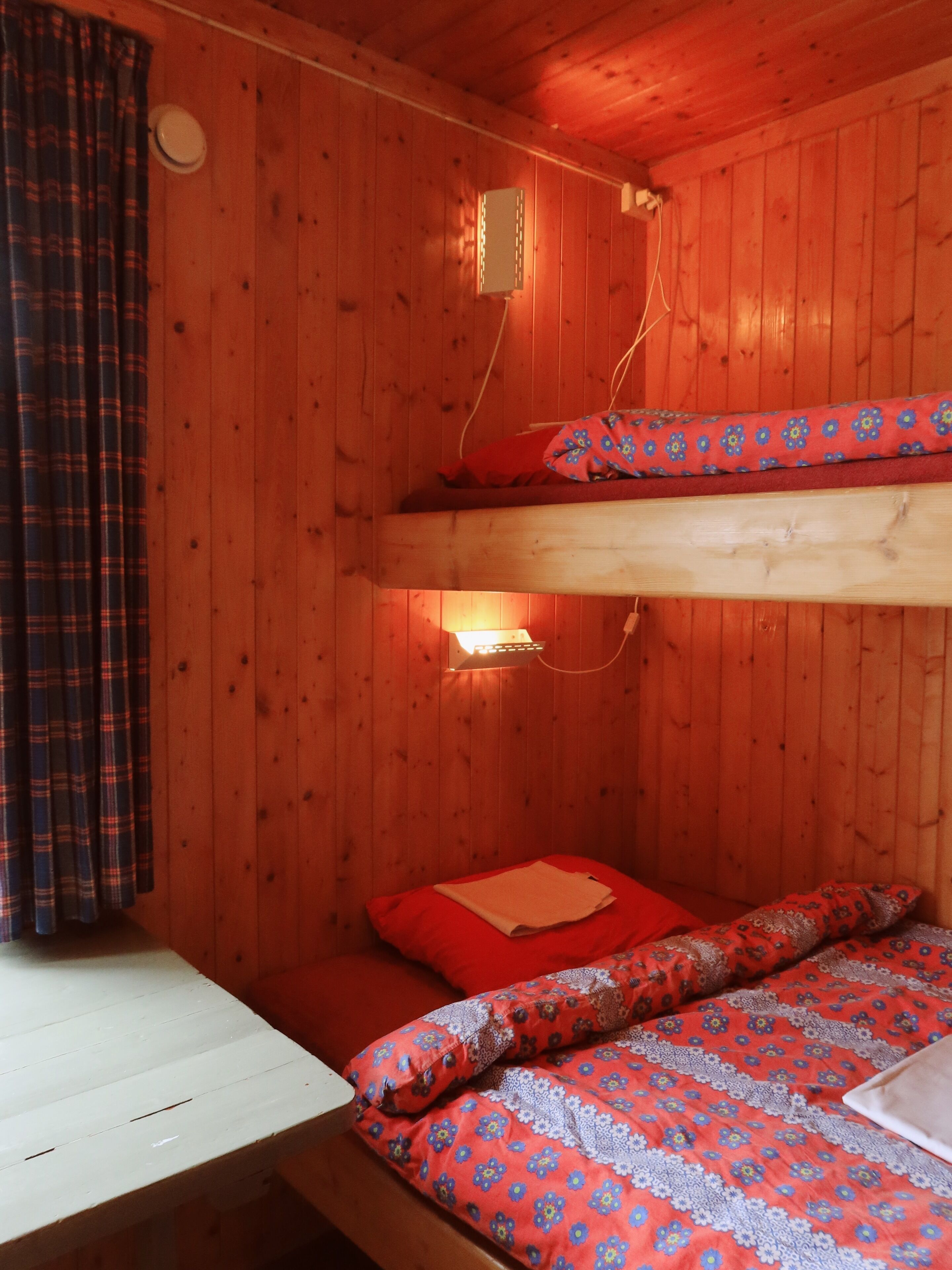 A wooden cabin room with bunk beds, a desk, and a window with curtains.