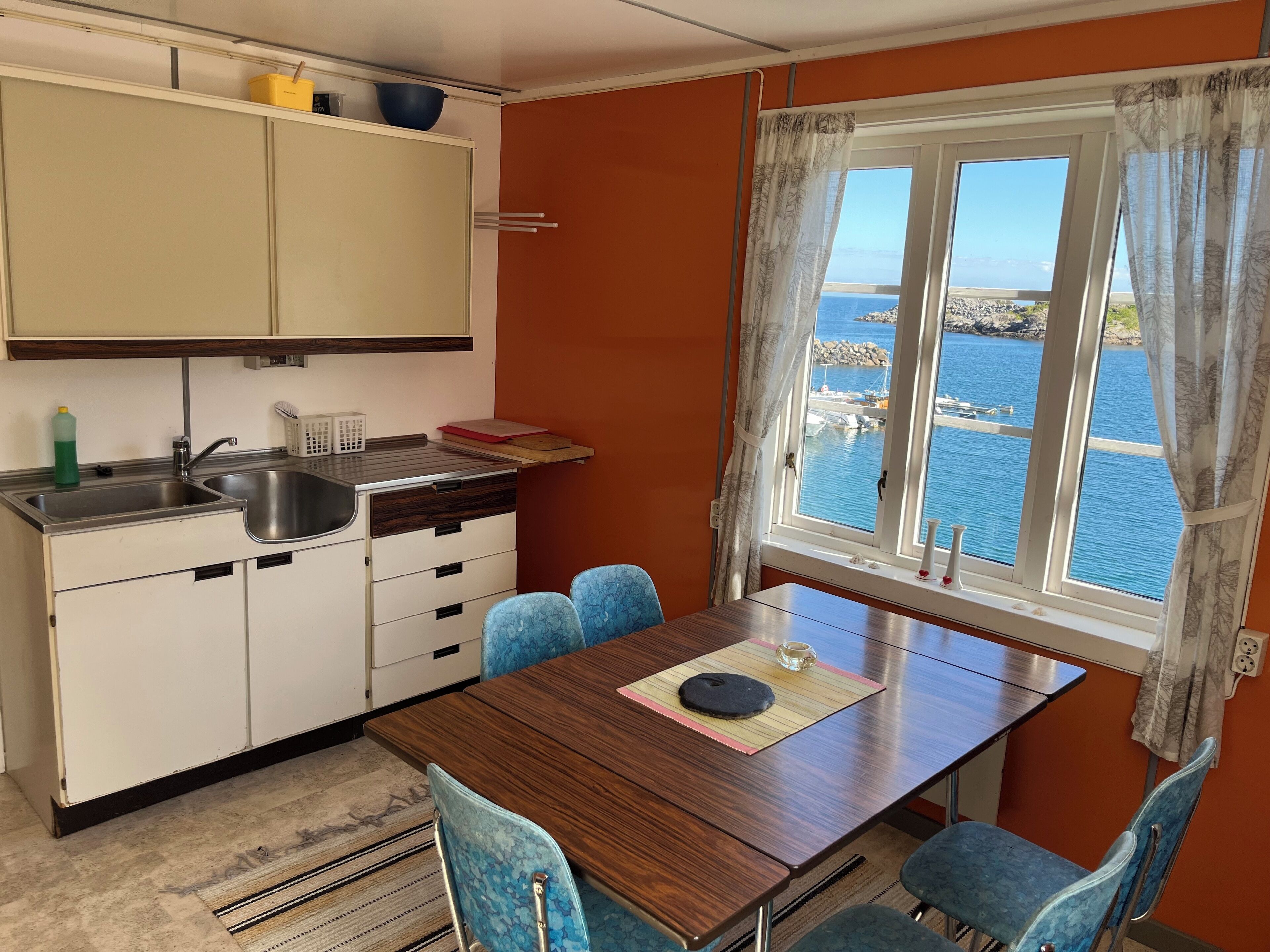 Une cuisine compacte comprenant un évier, des placards et un coin repas avec une table et des chaises. Une fenêtre offre une vue sur une étendue d’eau et une côte rocheuse.