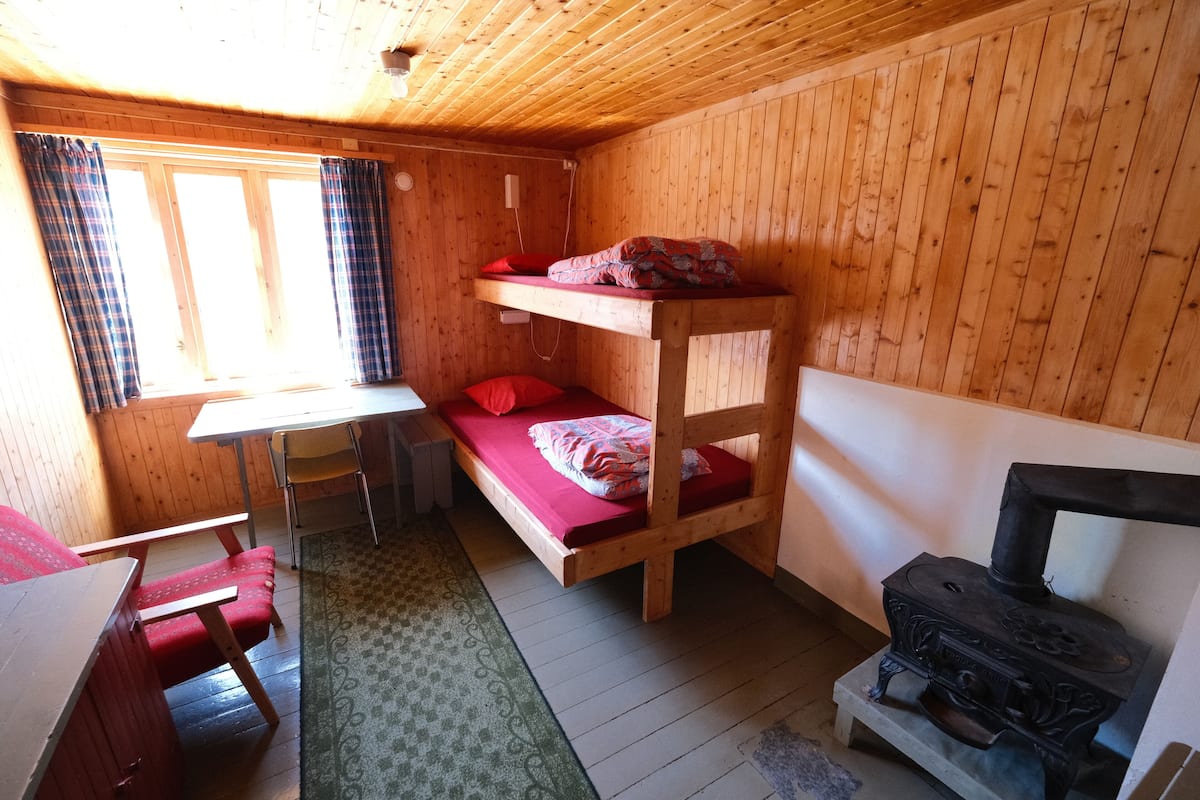 A wooden cabin room with bunk beds, a table, chairs, a stove, and a window with curtains.