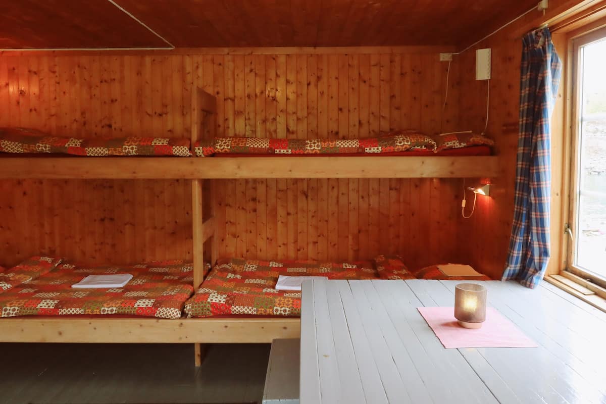 A cabin with two bunk beds, a wooden interior, a window with a curtain, and a small table with a candle.