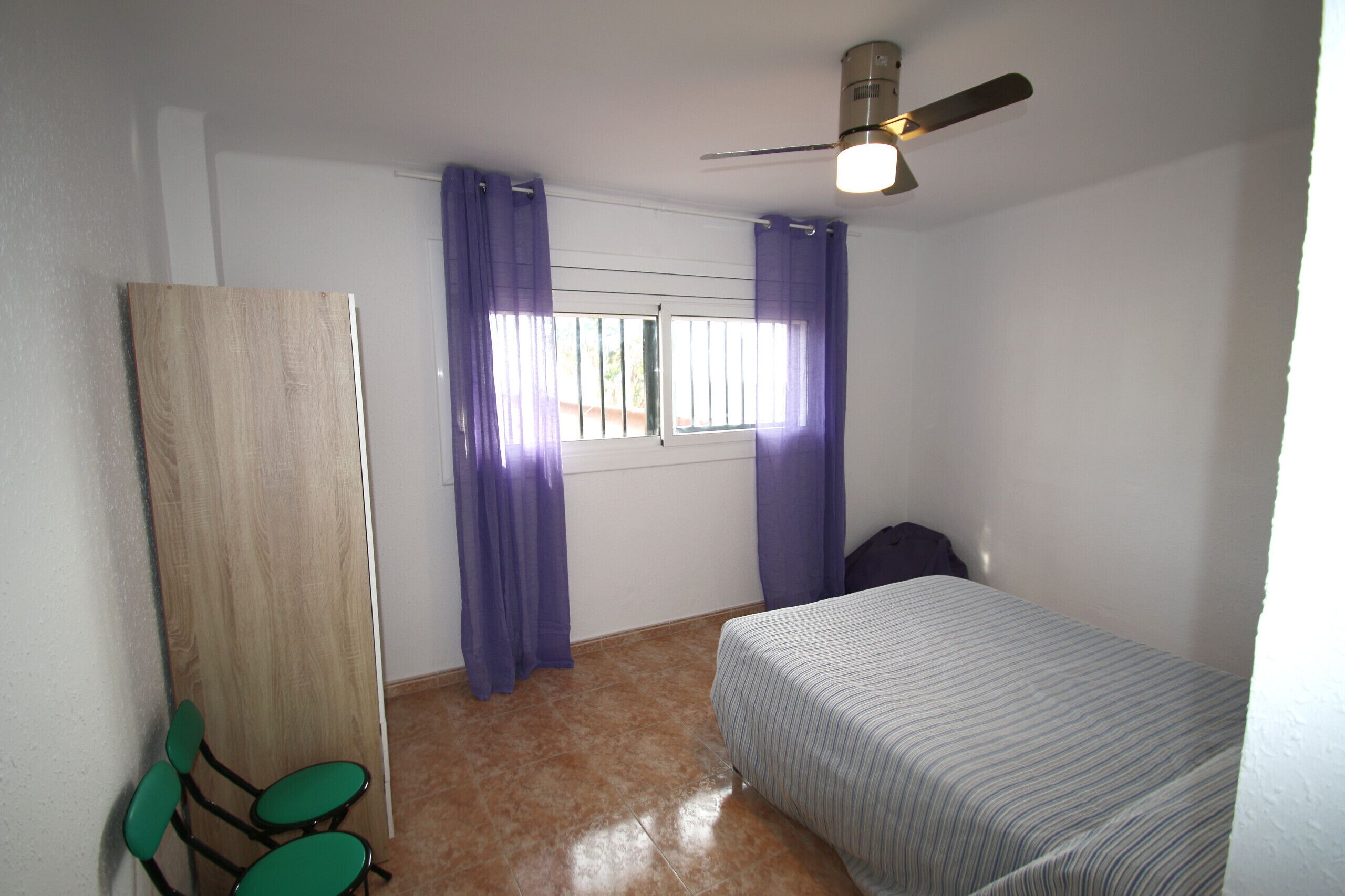A bedroom with a bed, a ceiling fan, a window with purple curtains, and a wooden door.