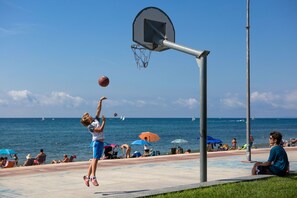 Un panier de basket installé sur une plage, offrant une vue dégagée sur l’océan et où l’on peut voir des gens se détendre sous des parasols.