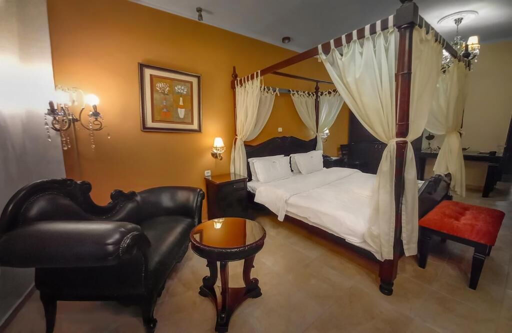 Una cama con dosel con sábanas blancas, un reposapiés rojo, un sillón de cuero negro y una pequeña mesa redonda con una vela.