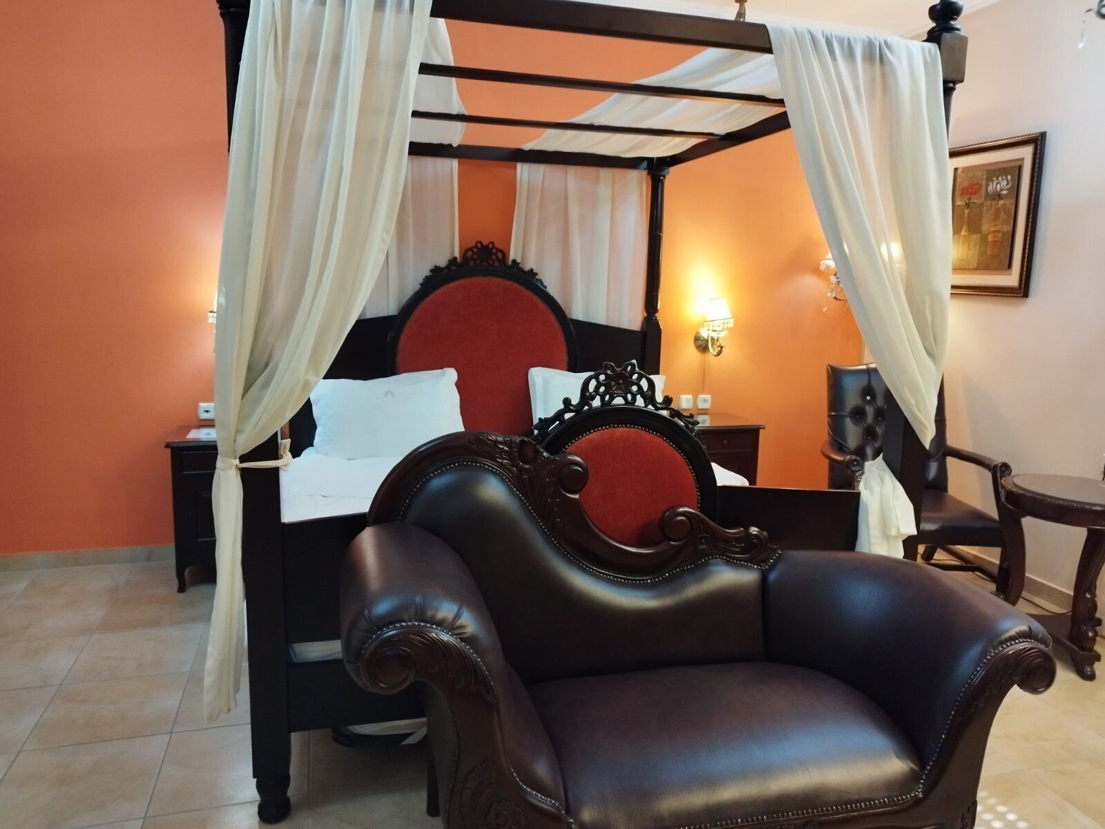 Una cama con dosel y cortinas blancas, un cabecero tapizado en rojo y un sillón de cuero negro.