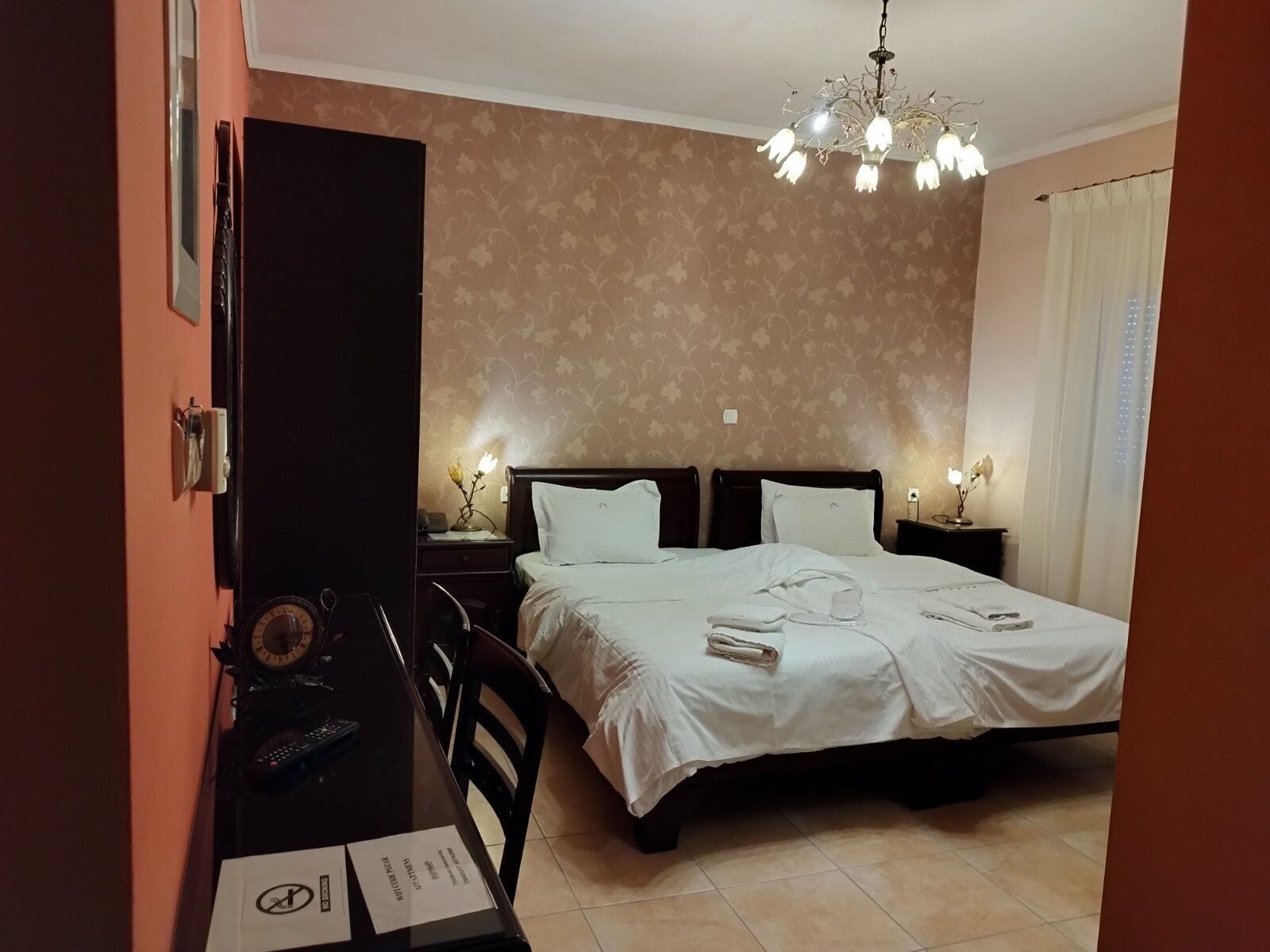 A hotel room with a bed, bedside tables, a desk with a chair, a clock, and a chandelier.
