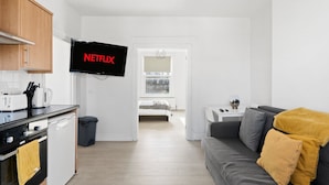 A modern kitchen with a built-in oven, a microwave, and a coffee maker. There is a TV mounted on the wall displaying the Netflix logo. The room has a grey sofa with a mustard throw blanket and a small white table with a remote control. - TTSA - Essential 1 Bedroom Apartment 2 - Croydon (Croydon)