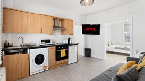 A modern kitchen with wooden cabinets, a washing machine, a microwave, and a TV displaying 'NETFLIX'. - TTSA - Essential 1 Bedroom Apartment 2 - Croydon (Croydon)