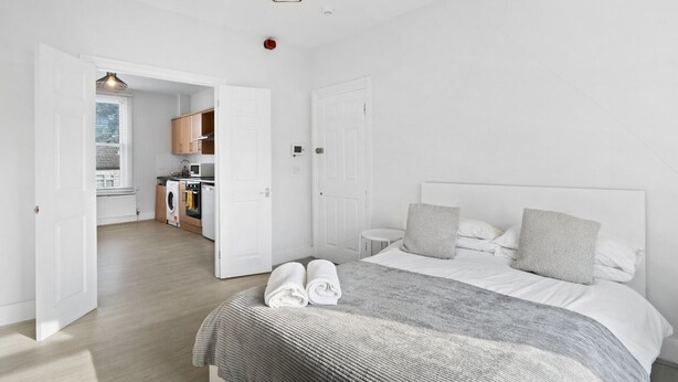 A bedroom with a bed, two pillows, and a bedside table. There is a bathroom with a toilet and sink. The room has white walls and a wooden floor. - TTSA - Essential 1 Bedroom Apartment 2 - Croydon (Croydon)