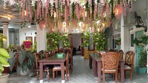 A dining area with wooden tables and chairs, surrounded by potted plants and hanging floral decorations.