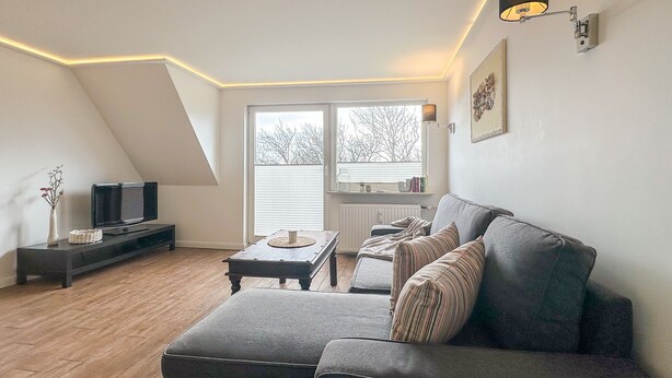 Ein modernes Loft-Apartment mit schrägem Dach, einem grauen Sofa, einem kleinen Couchtisch, einem Fernseher auf einem Schrank und einem großen Fenster mit Jalousien.