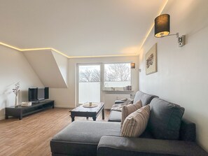 Un appartement moderne de style « loft » avec un plafond en pente, un canapé gris, une petite table basse, un téléviseur posé sur un meuble et une grande fenêtre avec des stores.