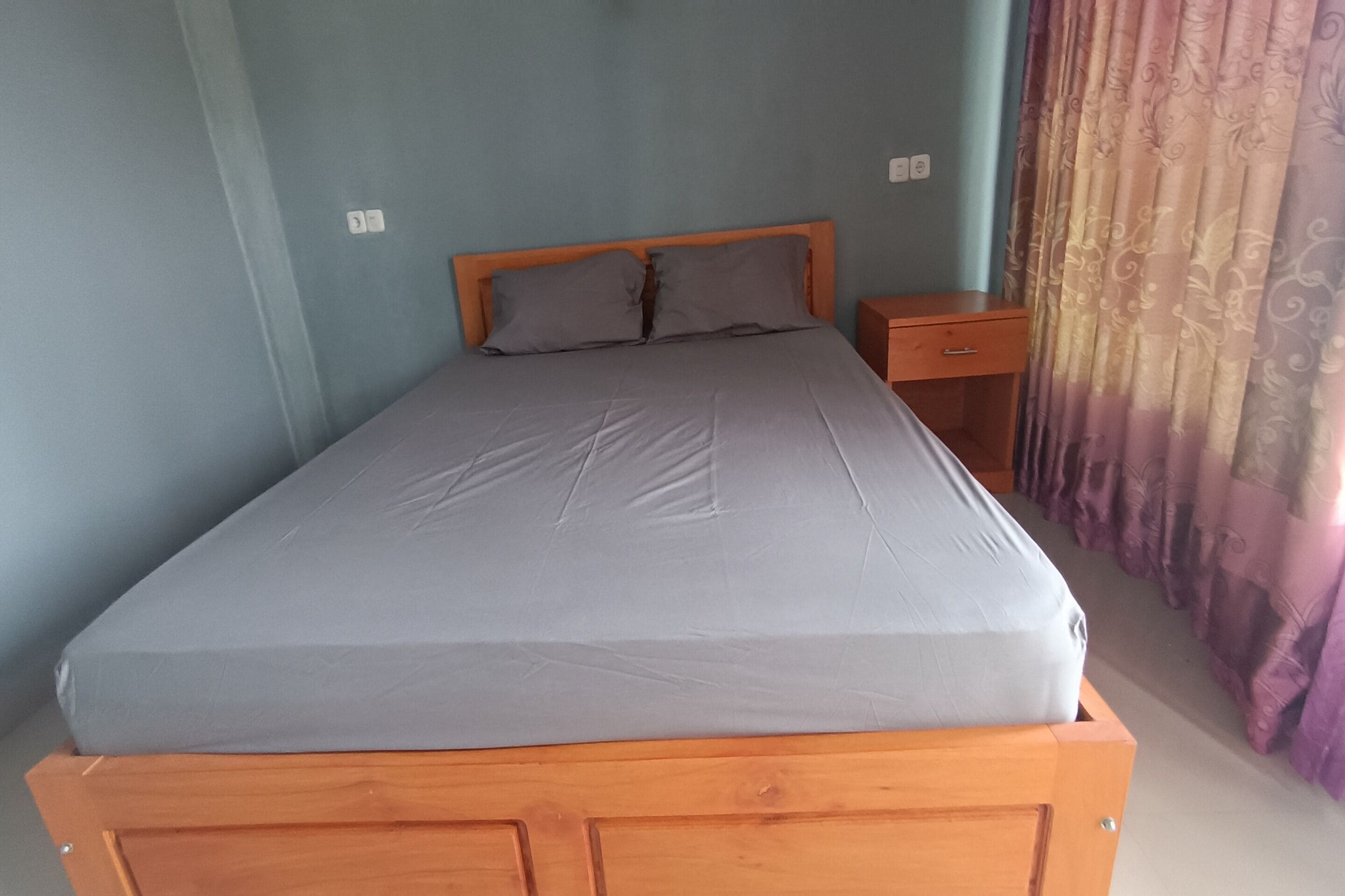 A single bed with a wooden headboard and a grey mattress in a room with light blue walls.