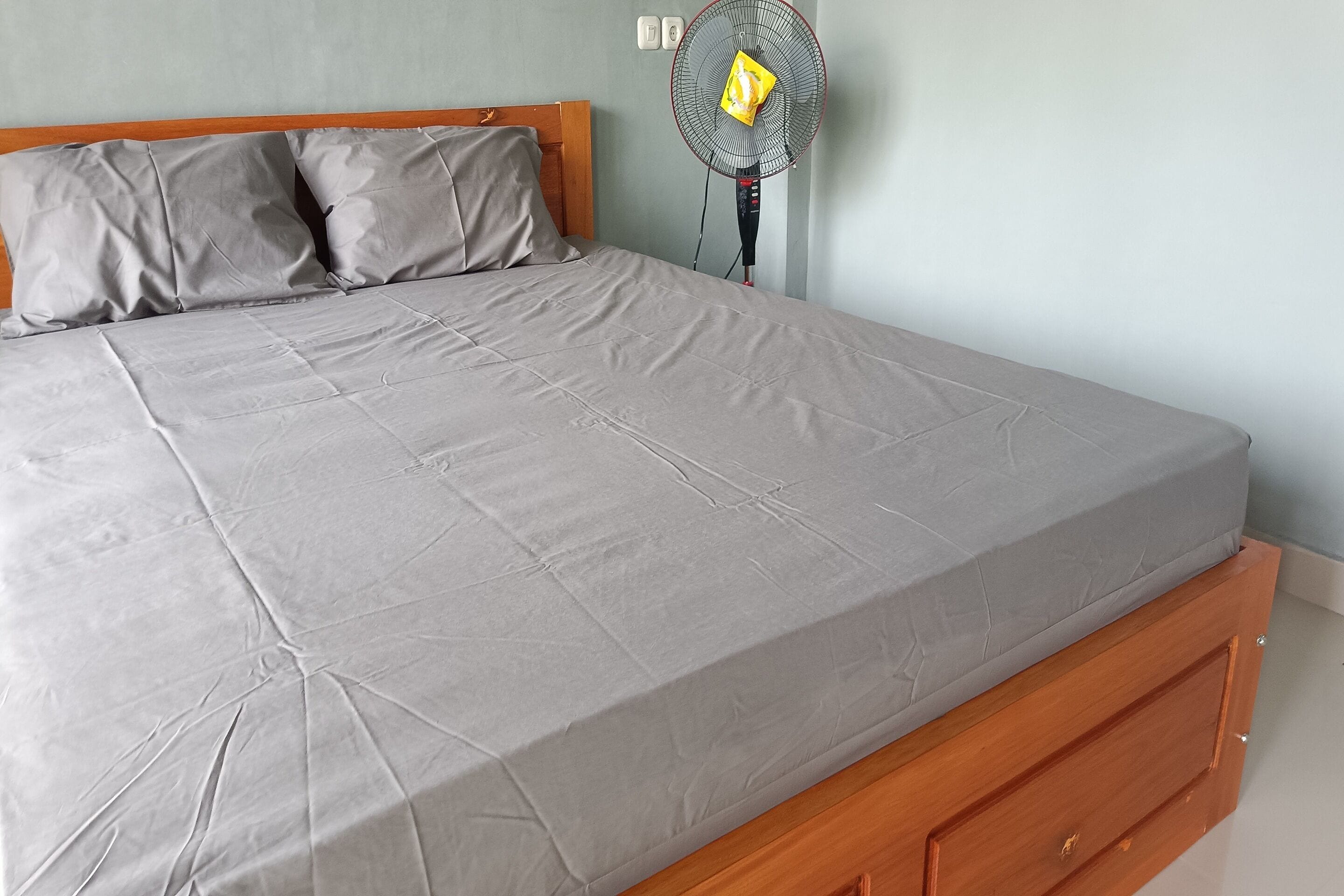 Una cama con colchón gris y cabecera de madera, un ventilador de pie y un interruptor de luz en la pared.