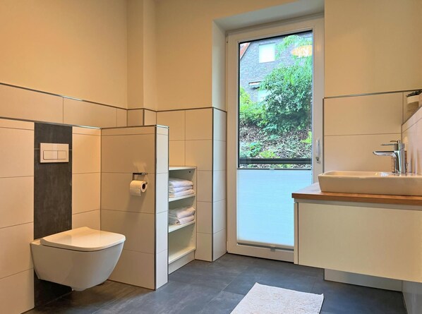 Een moderne badkamer met toilet, een douchecabine van glas, een wastafel met spiegel en een raam met uitzicht op groen.