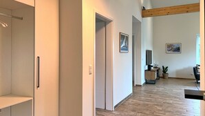 Ein modernes Interieur mit Holzboden, weißen Wänden und einem Holzbalken an der Decke. Eine Tür führt in einen anderen Raum, ein kleiner Tisch mit einer Pflanze und ein gerahmtes Bild an der Wand sind ebenfalls zu sehen.