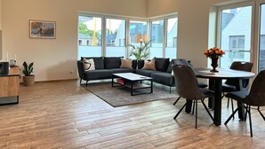 Ein geräumiges Wohnzimmer mit einer Sofagarnitur, einem Couchtisch und einem Esstisch mit Stühlen. Große Fenster bieten einen Ausblick nach draußen.