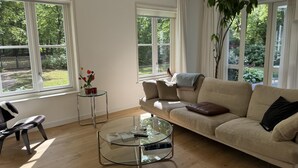 Een moderne woonkamer met een beige bank, een glazen salontafel en een bijzettafeltje met een bloemenvaas. Grote ramen bieden uitzicht op een tuin.