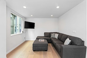 A modern living room with a sectional sofa, a flat-screen TV mounted on the wall, and a wooden floor.