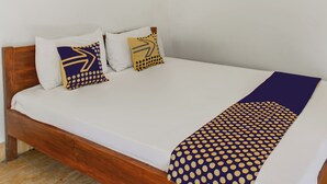 Una cama con colchón blanco, dos almohadas con diseños geométricos y una manta púrpura con estampado de lunares.