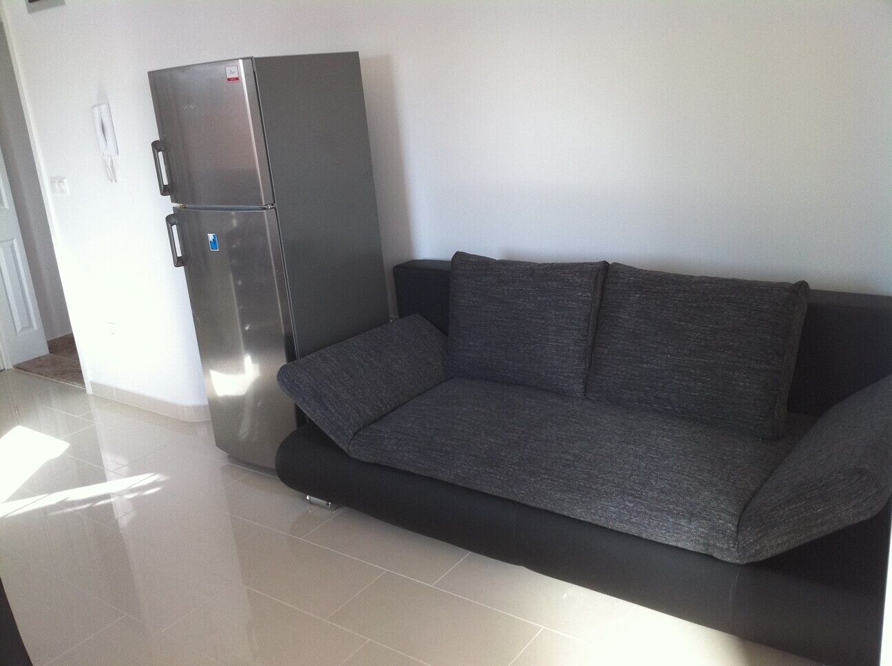 A modern living room with a grey sofa, a stainless steel refrigerator, and a tiled floor.