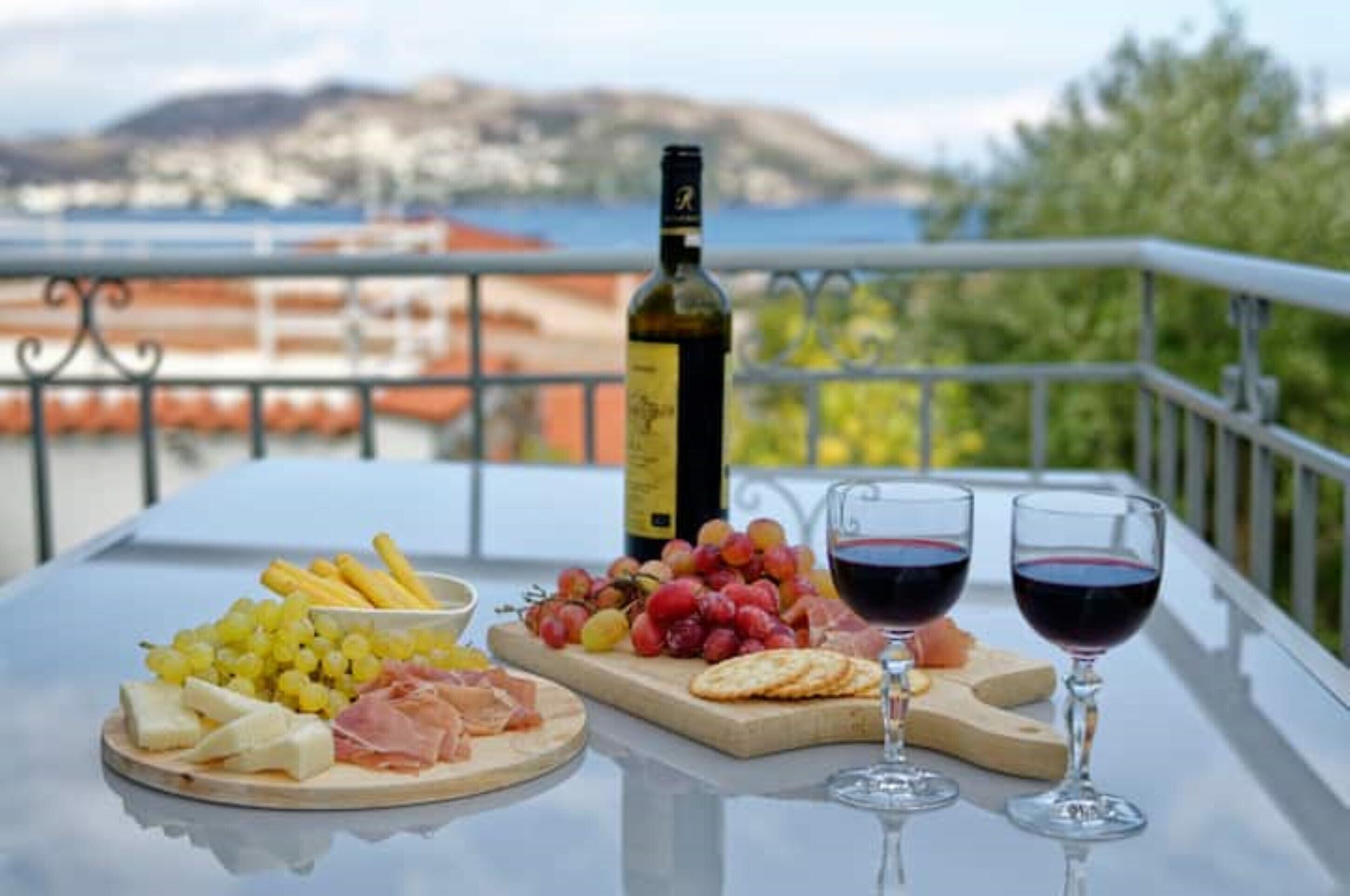 A table with a bottle of wine, glasses, and a variety of cheeses and fruits on a balcony with a scenic view.