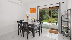 Un coin repas avec une table en verre, des chaises noires, un climatiseur blanc et une vue sur un jardin, visible à travers une porte-fenêtre coulissante.
