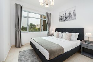A bedroom with a bed, curtains, a nightstand, a lamp, a painting, and a view through the window. - Lux BnB Greens & Views Lake View  (Dubai)