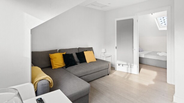 Un salon moderne et minimaliste, avec un canapé d’angle gris, une petite table d’appoint blanche et une table basse en verre. Une porte donne accès à une chambre à coucher équipée d’un lit et d’une fenêtre.