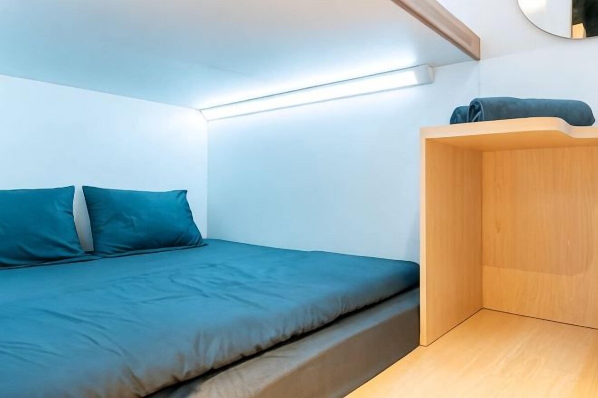 Una habitación pequeña y funcional con una cama individual, un banco de madera y un espejo montado en la pared.