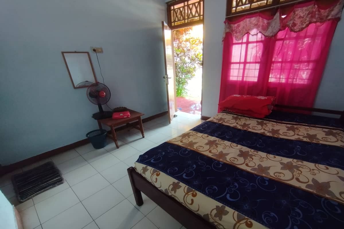 A bedroom with a bed, a standing fan, a small table, and a window with curtains.