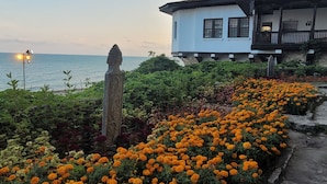 A coastal building with a white facade and a green spire, surrounded by a garden of orange flowers and a stone pathway. - 3-room new apartment,  5-min walk to 3 sandy beaches  (Topola Village)