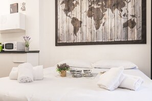 Ein ordentlich bezogenes Bett mit weißen Leinen und Handtüchern, eine kleine Pflanze und zwei Tassen auf einem Tablett. Über dem Bett hängt ein Wandbild einer Weltkarte.