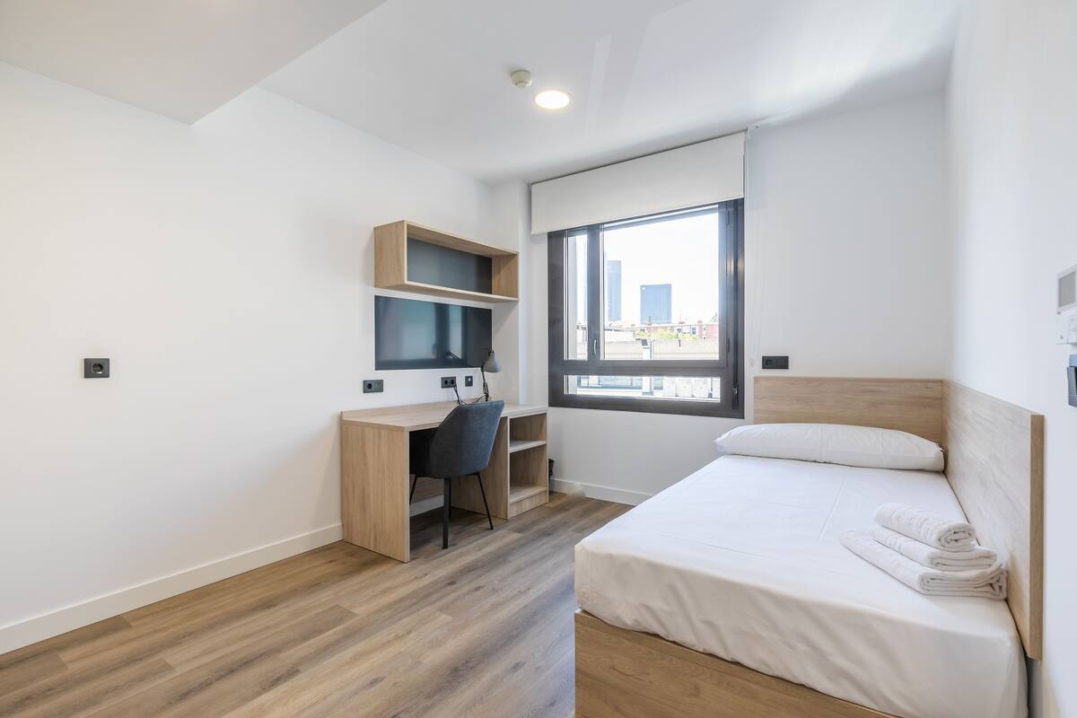 Una habitación de hotel moderna con un escritorio de madera, una cama con ropa blanca y un ventanal con vistas a la ciudad.