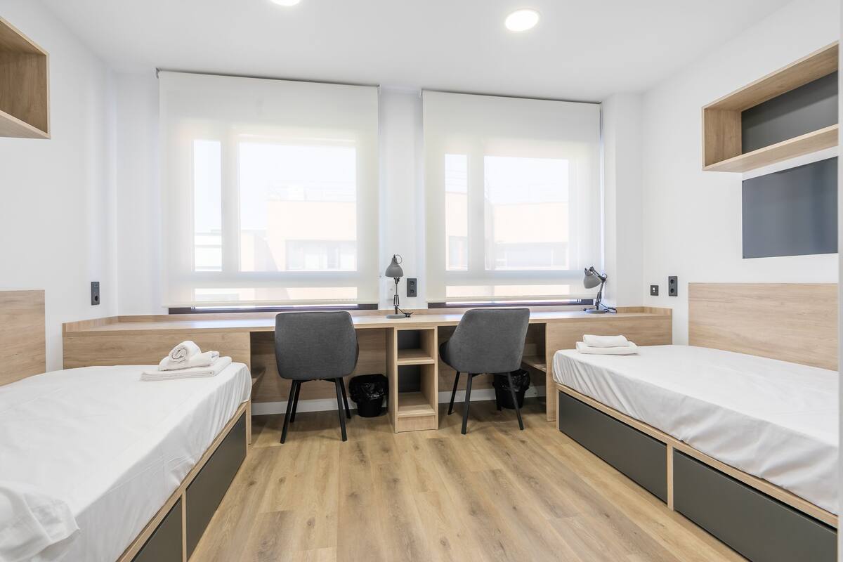 Habitación de hotel moderna con dos camas individuales, un escritorio, dos sillas y un ventanal amplio.