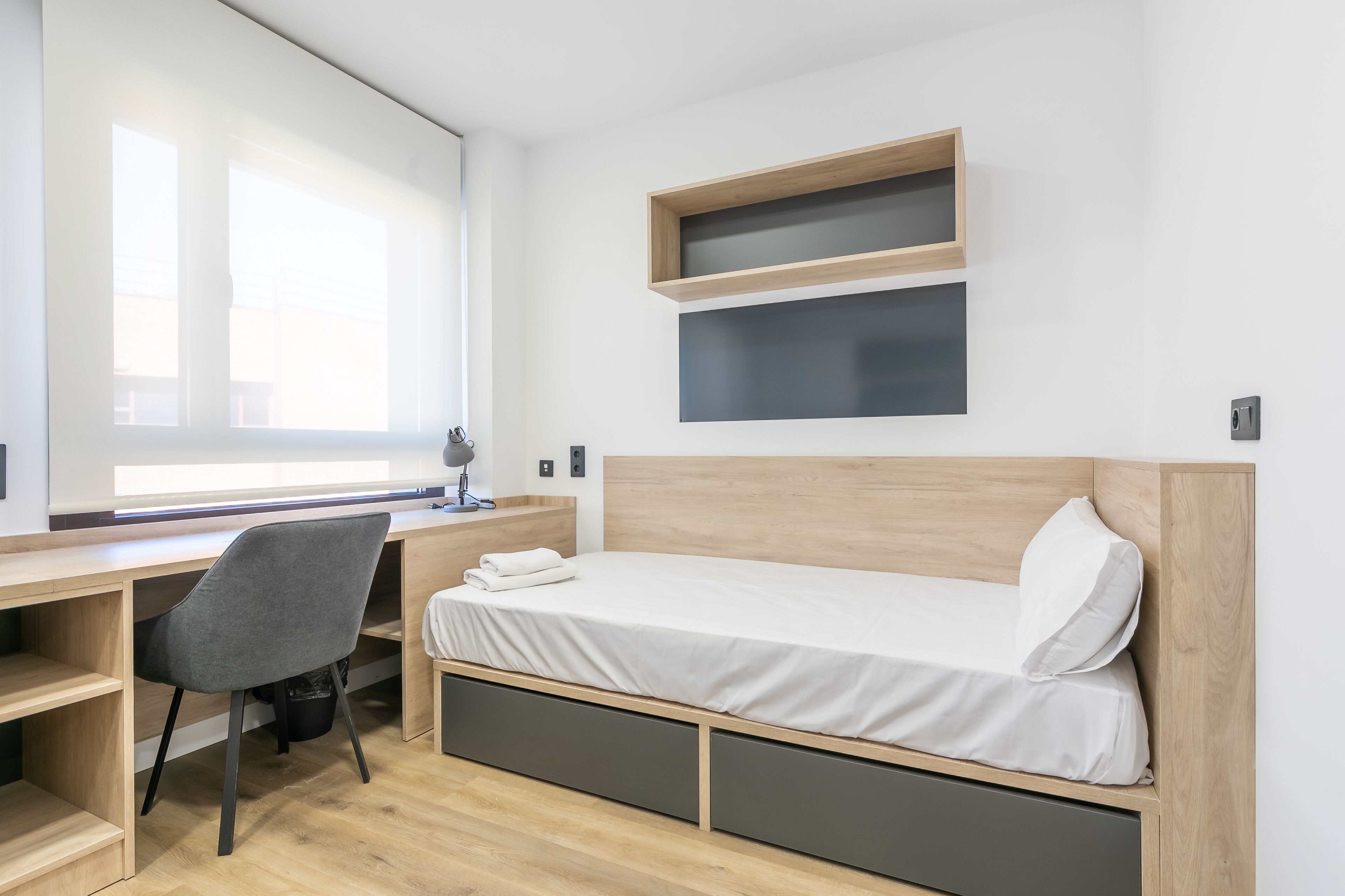 A modern bedroom with a wooden bed, a desk, and a chair. There are two shelves above the bed and a window providing natural light.