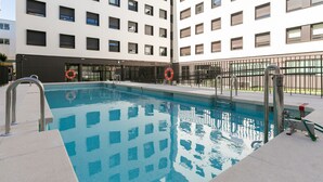 A swimming pool with a metal ladder and lifebuoys, surrounded by a fence and a building with multiple windows. - Micampus Estrada 11 (Madrid)