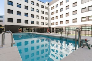 Ein Schwimmbecken mit Metallleiter und Rettungsringen, umgeben von einem Zaun und einem Gebäude mit zahlreichen Fenstern.
