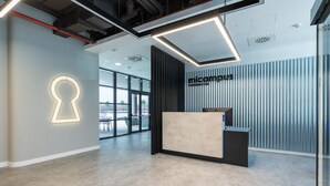 A modern reception area with a minimalist design, featuring a reception desk, a key-shaped light installation, and a view of the outdoors through large windows. - Micampus Estrada 11 (Madrid)