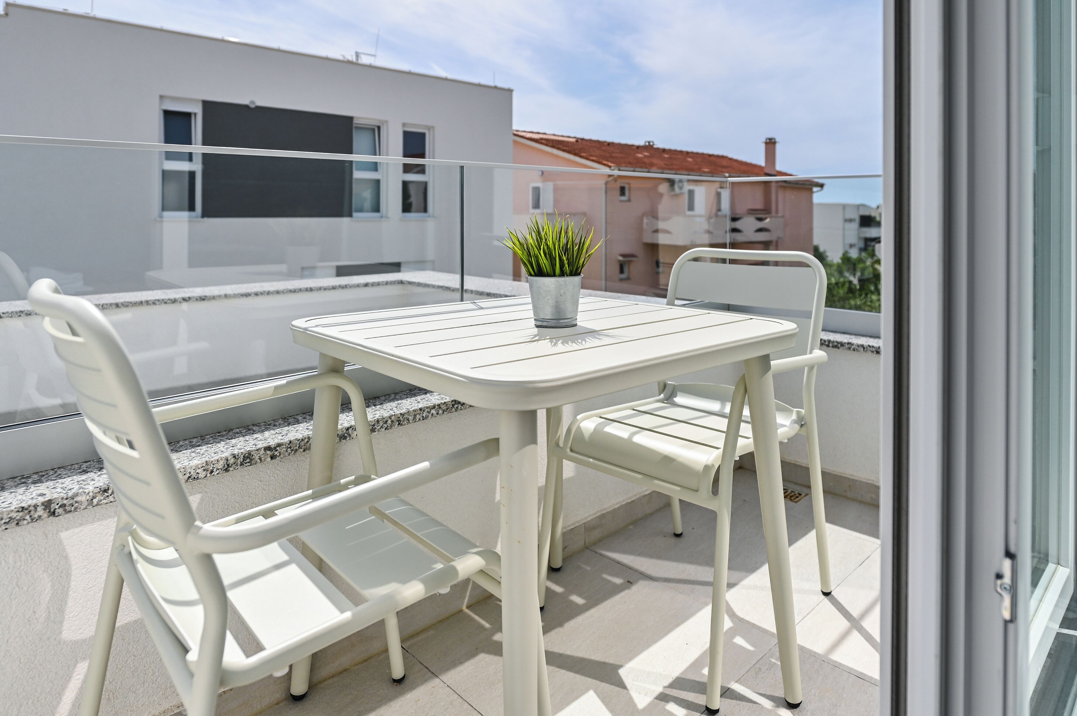 Een balkon met een witte tafel en stoelen, een bloempot en uitzicht op omliggende gebouwen.