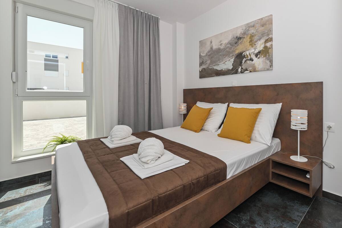 A hotel room with a bed, a nightstand, a lamp, a window with curtains, and a painting on the wall.