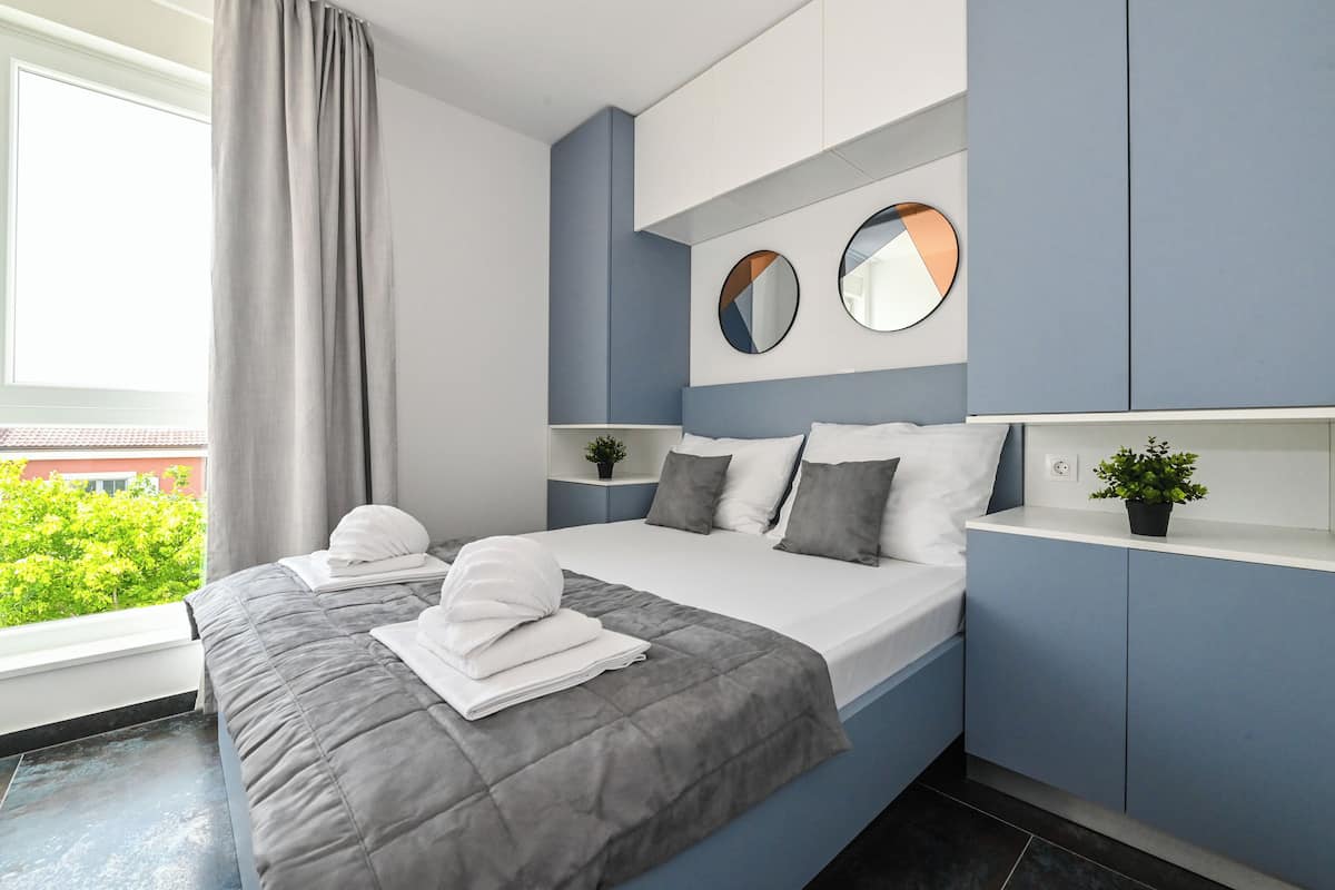 A modern bedroom with a bed, two bedside tables, a large window, and a view of greenery outside.
