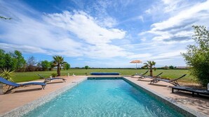 Una piscina rectangular con agua azul cristalina, rodeada por una terraza de madera. Hay sillas de descanso y una palmera en la terraza.