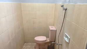 Une petite salle de bain carrelée, avec des toilettes, un seau rose et une pomme de douche.